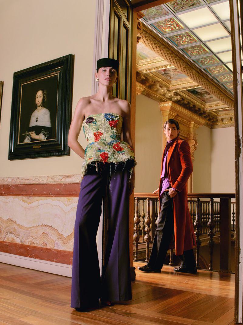 Model wearing a strapless floral top and wide-leg purple trousers stands in an ornate classical hallway beside a framed painting.