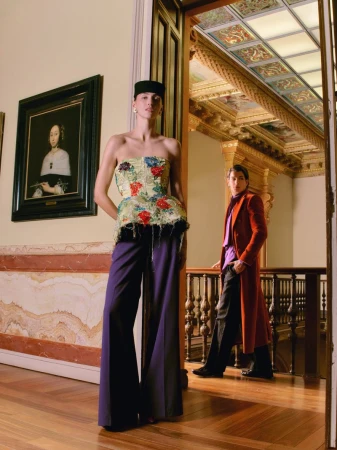 Model wearing a strapless floral top and wide-leg purple trousers stands in an ornate classical hallway beside a framed painting.