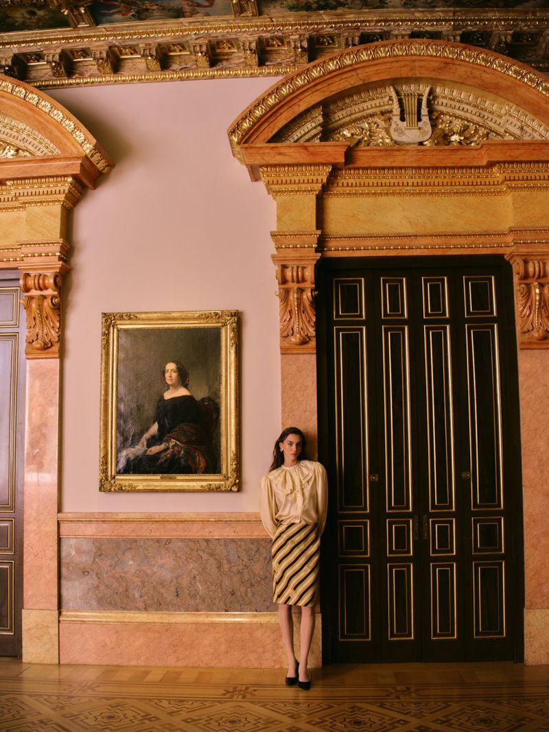 Model in a cream blouse and striped skirt standing in an ornate room beside a tall black double door and framed painting.