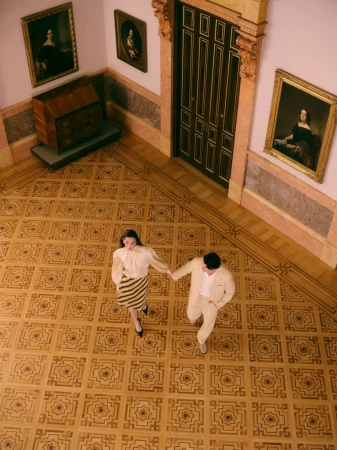 High-angle shot of a man and woman holding hands walking across an ornate parquet floor inside a classical gallery.