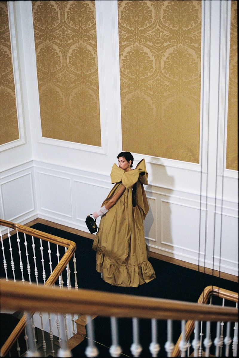 A woman in a voluminous gold gown poses on a dark staircase against gold damask patterned walls framed by white moulding.