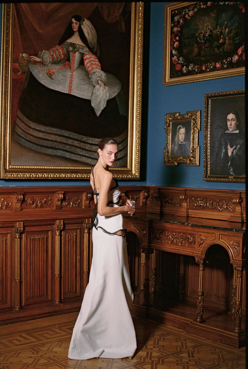 Model in a floor-length white evening gown looking over her shoulder in a classic gallery room with framed paintings.