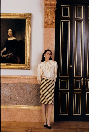 Model wearing a ruffled cream blouse and diagonal striped skirt standing between an oil painting and a tall black paneled door.