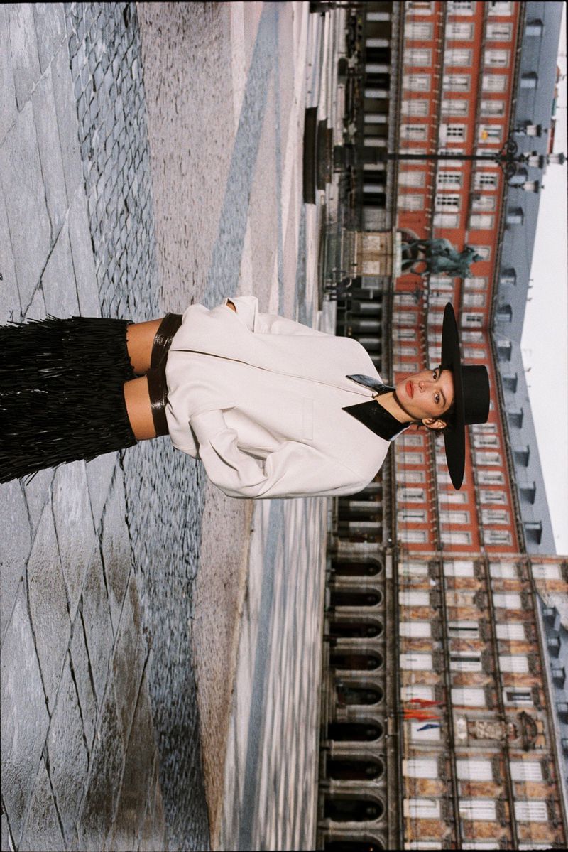 Model wearing a wide-brimmed black hat, white jacket, and black fringe skirt standing in Plaza Mayor in Madrid.