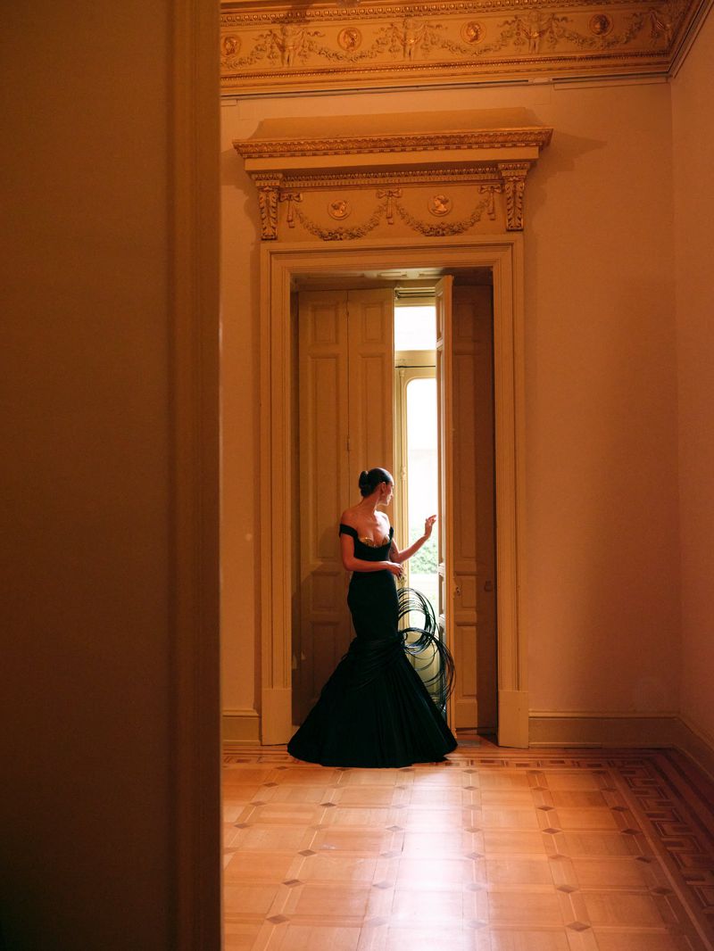 A model wearing a black off-the-shoulder evening gown stands in an ornate doorway with parquet flooring looking through double doors.
