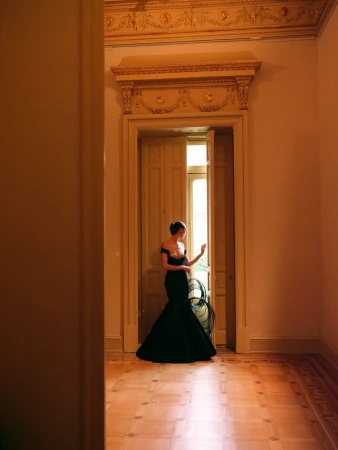 A model wearing a black off-the-shoulder evening gown stands in an ornate doorway with parquet flooring looking through double doors.