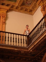 A woman in an asymmetrical white dress standing on an ornate wooden staircase landing beneath a decorated coffered ceiling.