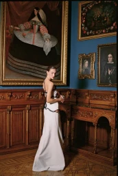 Model in a floor-length white evening gown looking over her shoulder in a classic gallery room with framed paintings.