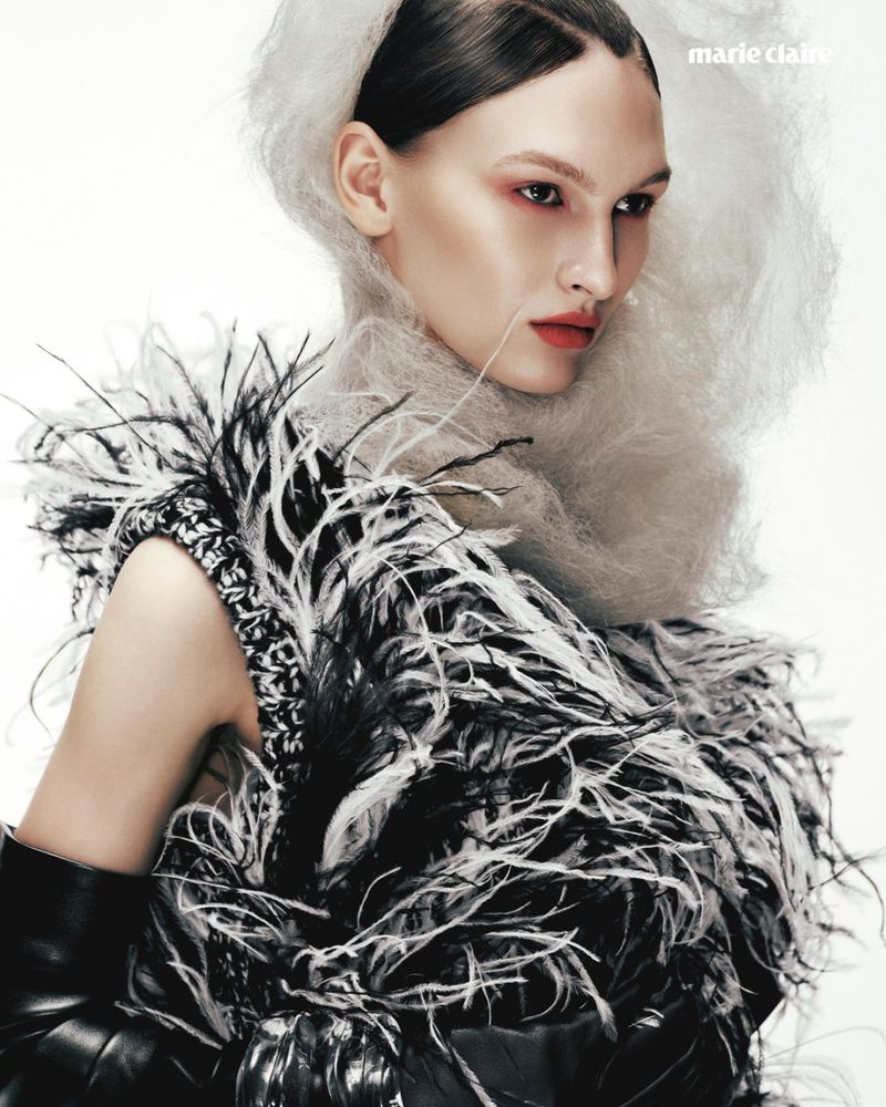Astrid Baauer wearing an avant-garde black and white feathered garment and white mesh headdress against a white studio background.
