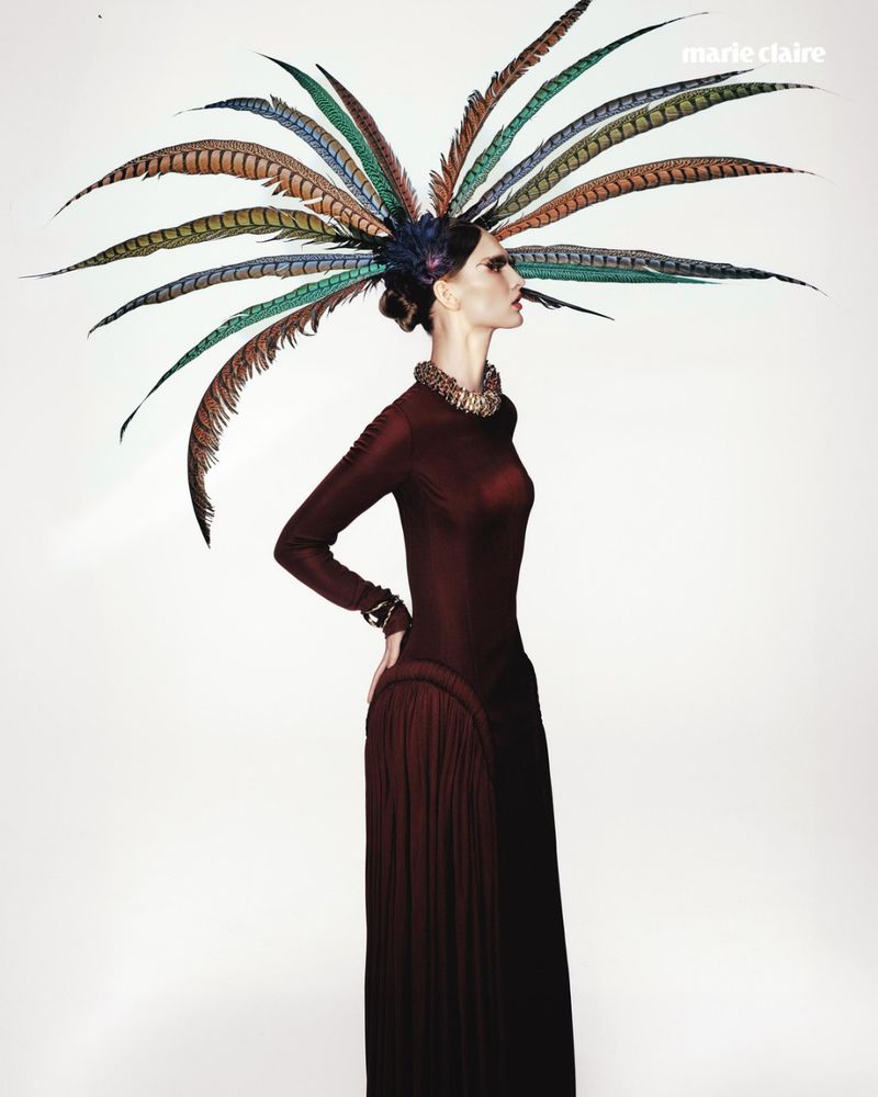 Model Astrid Baauer in profile wearing a large radiating feather headdress and a long-sleeve burgundy dress on a white background.