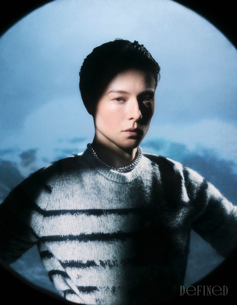 Portrait of Emma D'Arcy wearing a striped knit sweater and silver necklace inside a circular vignette frame against a blue sky background.