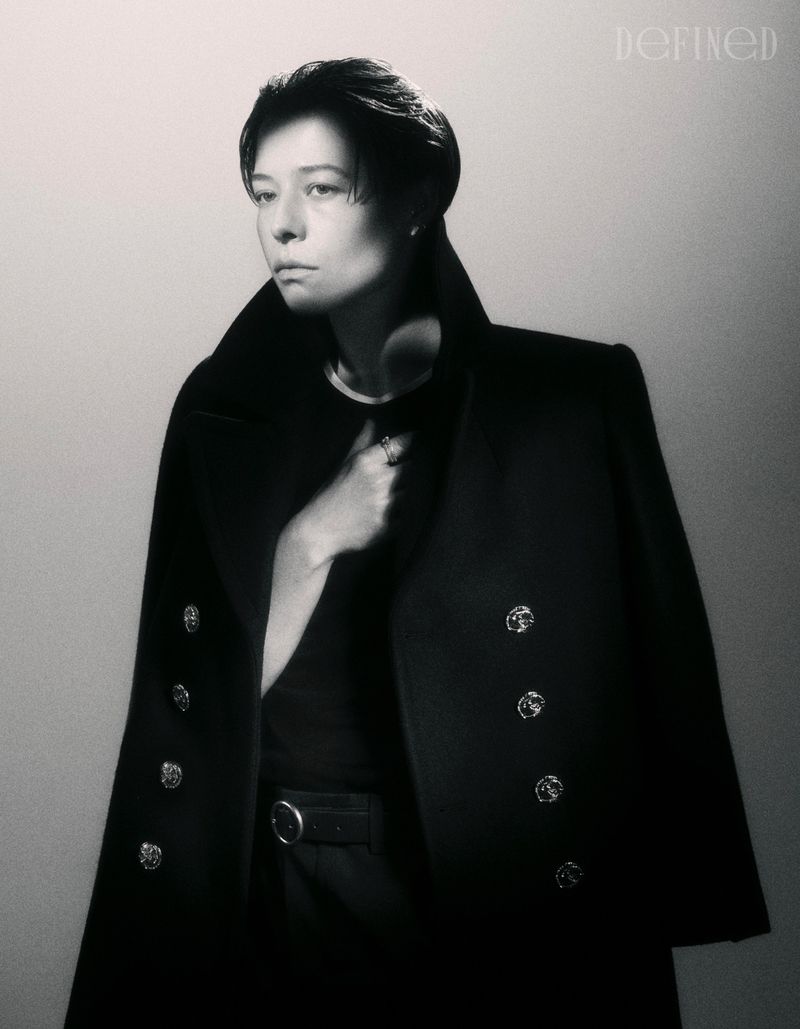 Emma D'Arcy posing in a tailored black double-breasted overcoat against a neutral studio backdrop for DEFINED Magazine.
