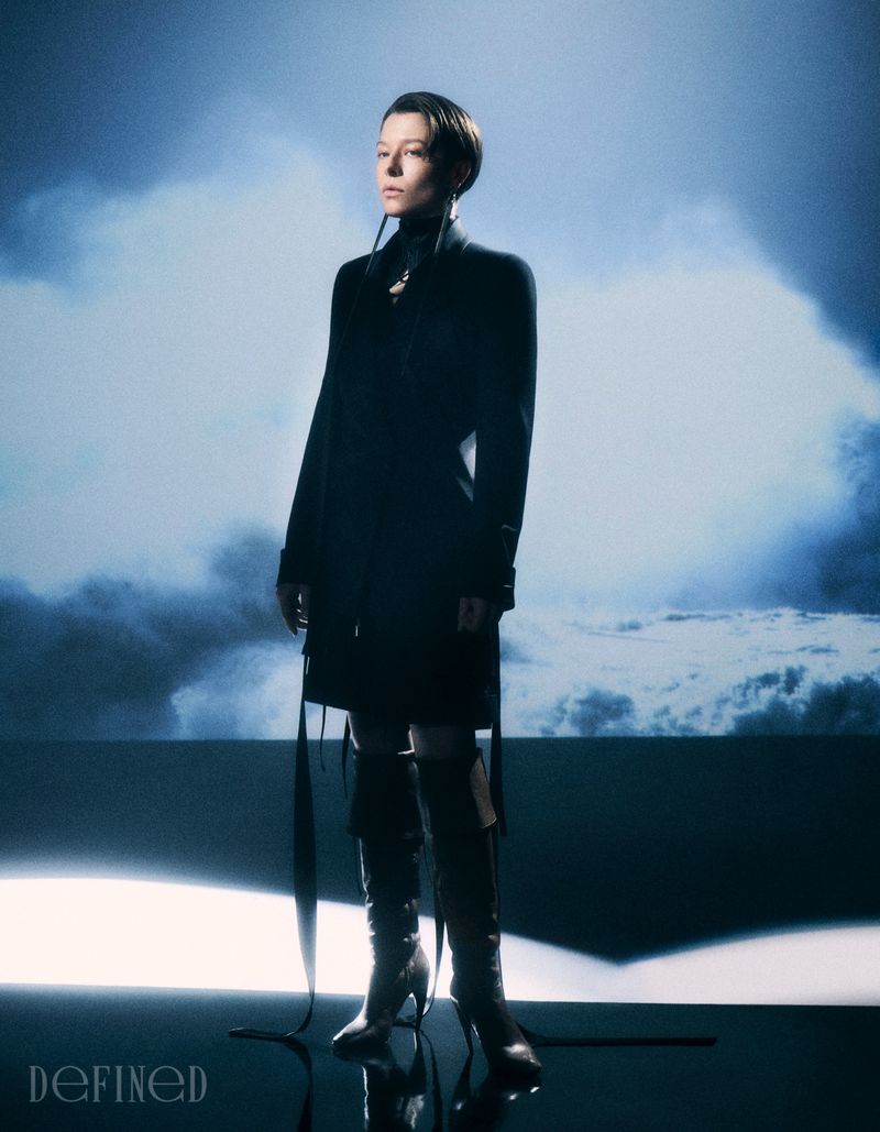 Emma D'Arcy wearing a black coat and tall boots against a blue cloud backdrop for DEFINED Magazine