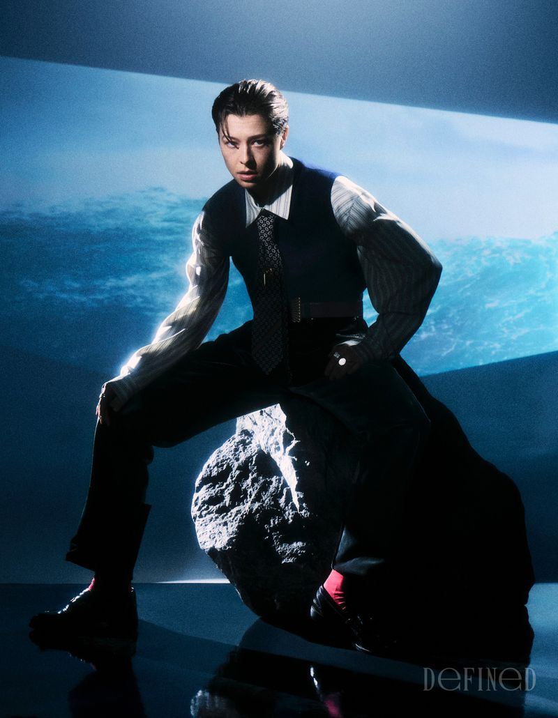 Actor Emma D'Arcy posing seated on a large rock wearing a dark waistcoat, tie, and striped shirt for DEFINED Magazine.