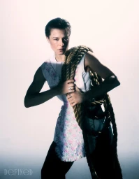 Emma D'Arcy posing with a thick coiled rope over their shoulder in a white floral tunic against a white backdrop.