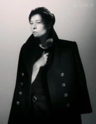 Emma D'Arcy posing in a tailored black double-breasted overcoat against a neutral studio backdrop for DEFINED Magazine.