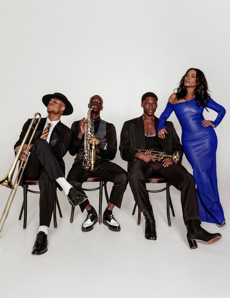 Nia Long in a royal blue gown standing beside three men in black suits holding brass instruments against a white studio backdrop.