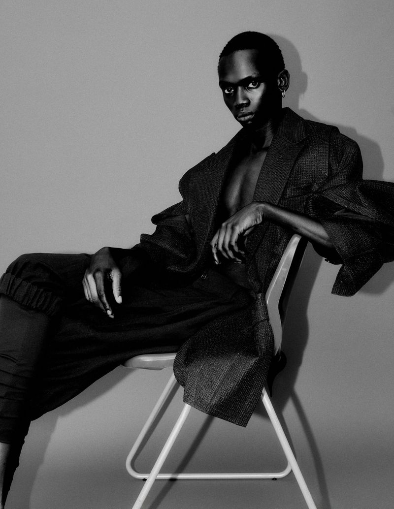 Model Bukwop Kir seated in an oversized textured coat on a minimalist chair against a studio backdrop in black and white.
