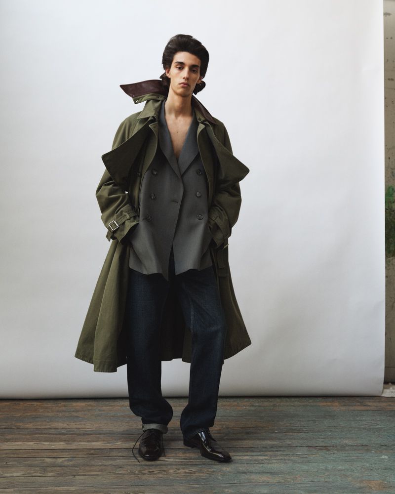 Model wearing an oversized olive green trench coat, grey blazer, and dark trousers against a white studio backdrop.