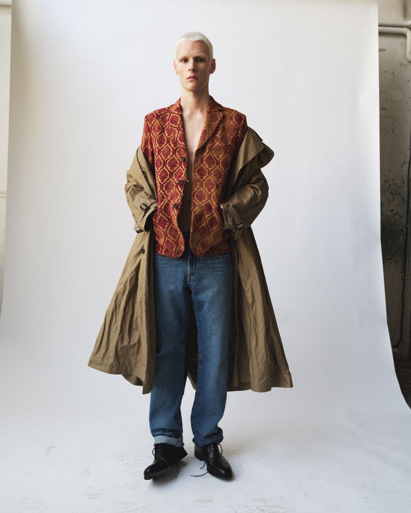 Model with bleached hair wearing a patterned red jacket and khaki coat over blue jeans against a white seamless background