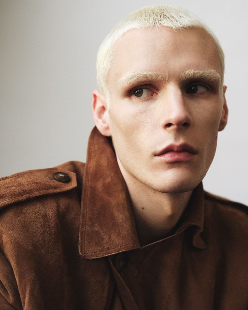 Close-up portrait of a man with bleached blonde hair wearing a brown suede jacket against a plain neutral background.