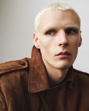 Close-up portrait of a man with bleached blonde hair wearing a brown suede jacket against a plain neutral background.