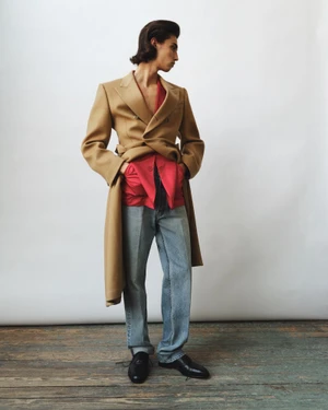 Male model posing in a camel coat, red shirt, and light wash denim jeans on a wooden floor in front of a white studio backdrop.