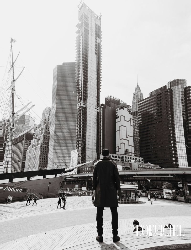 Model viewed from behind in a dark overcoat facing towering city skyscrapers and a masted ship in black and white.