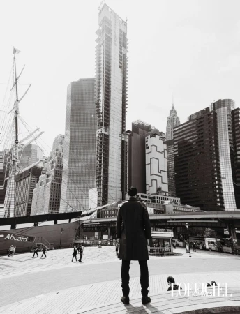 Model viewed from behind in a dark overcoat facing towering city skyscrapers and a masted ship in black and white.