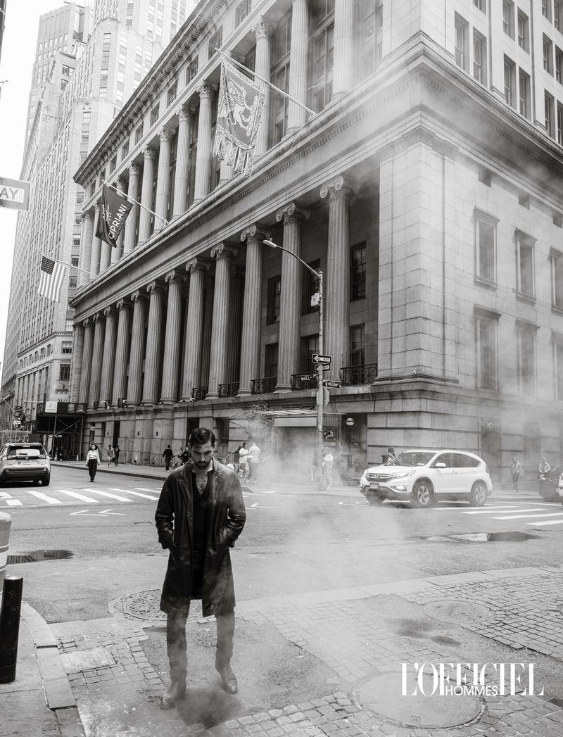 Black and white editorial photo of a man in a long coat standing amid rising street steam in front of a historic colonnaded building.