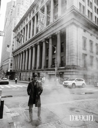 Black and white editorial photo of a man in a long coat standing amid rising street steam in front of a historic colonnaded building.
