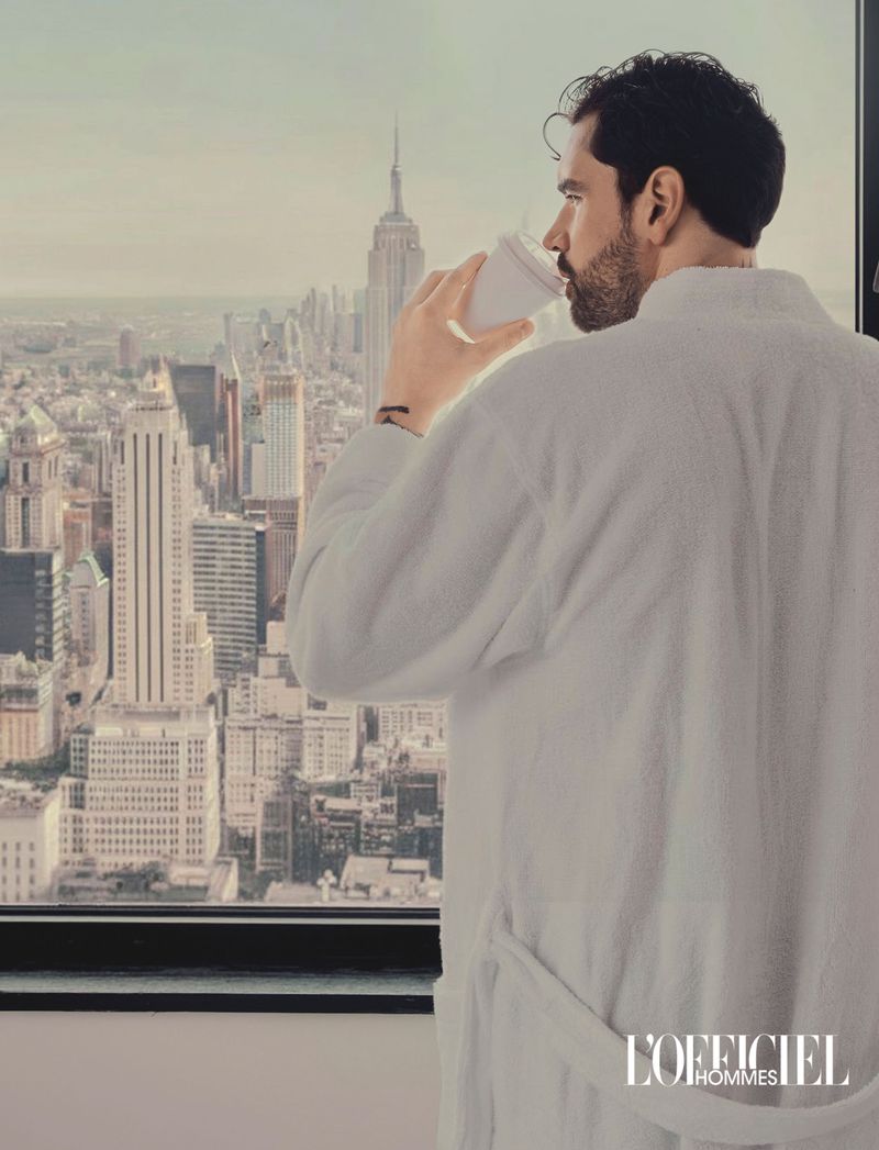 Man in white bathrobe drinking from a cup by a high-rise window overlooking a city skyline with skyscrapers.
