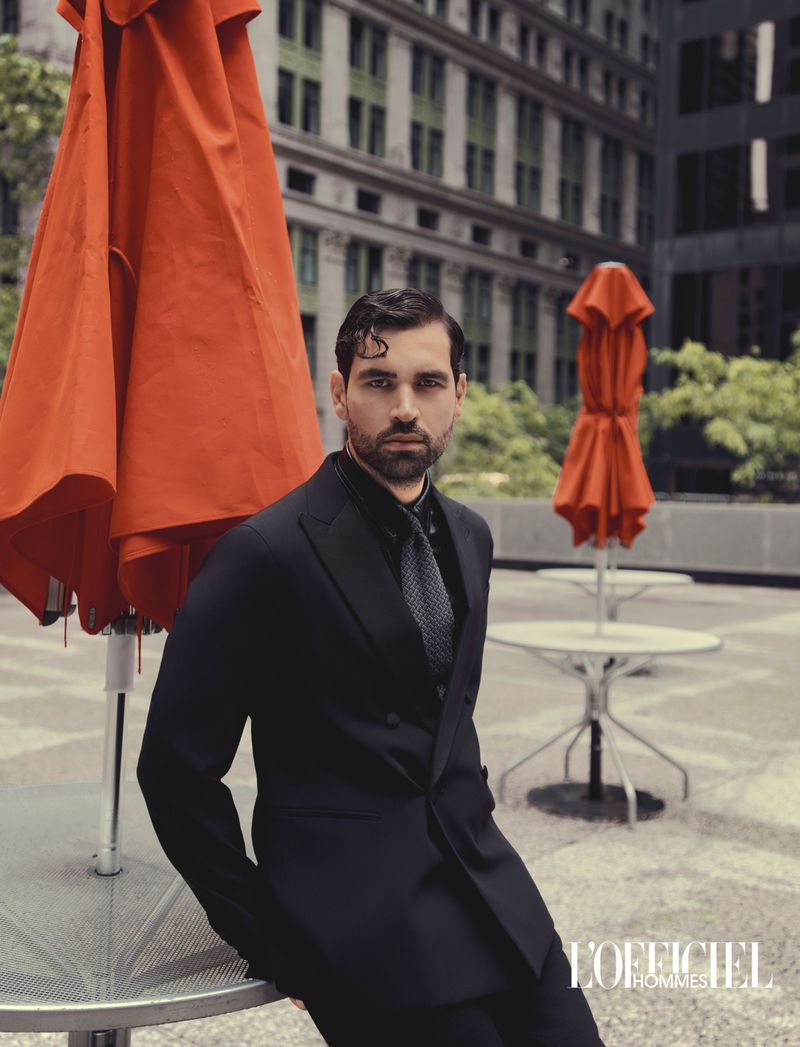 Model Emanuele Antonio Panico wearing a black double-breasted suit next to folded orange umbrellas in an urban plaza.