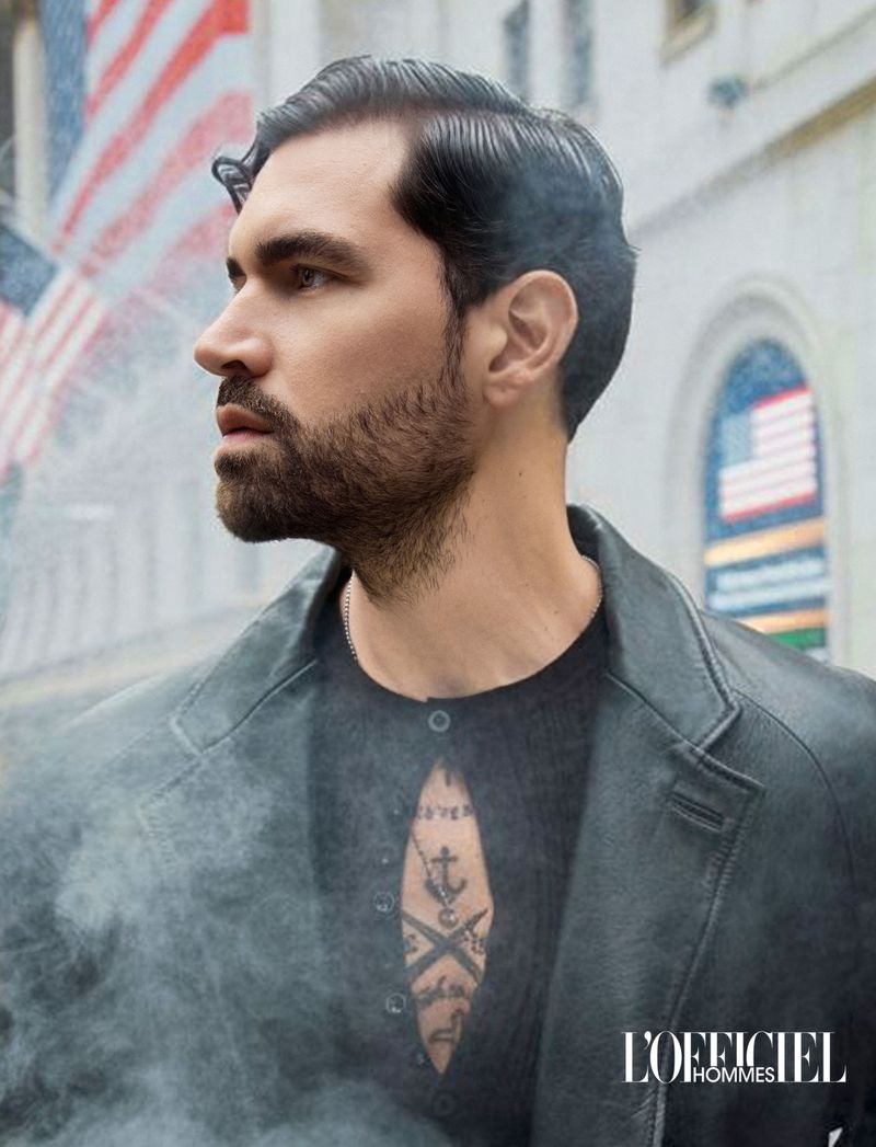 Profile portrait of a bearded man with a chest tattoo wearing a dark leather jacket in a misty urban street setting.