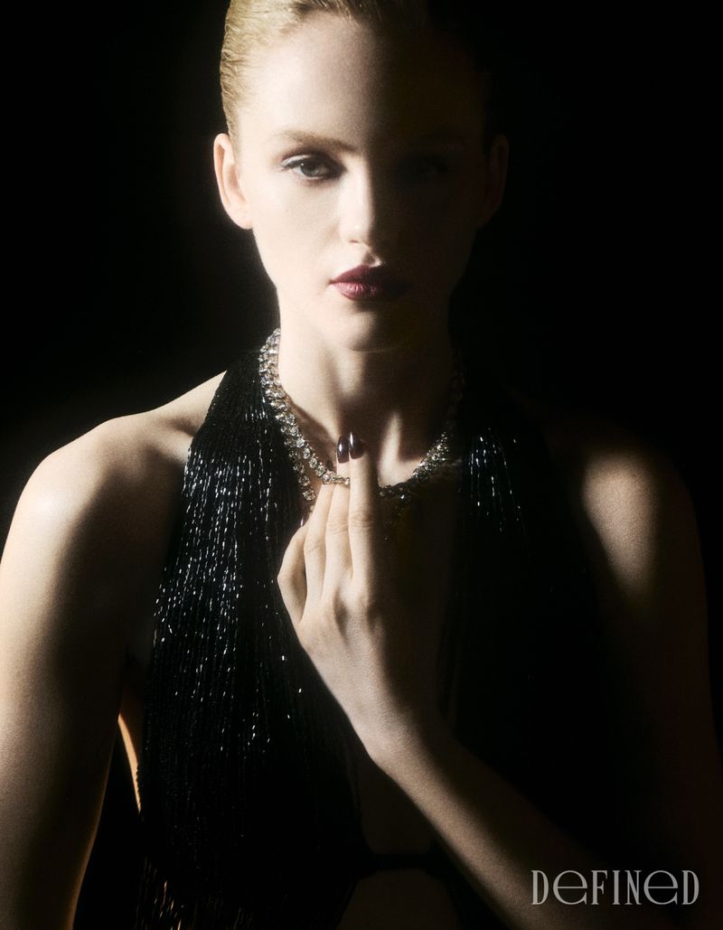 Emma Laird wearing a sparkling necklace and black halter fringe dress in a low-key portrait against a black background.