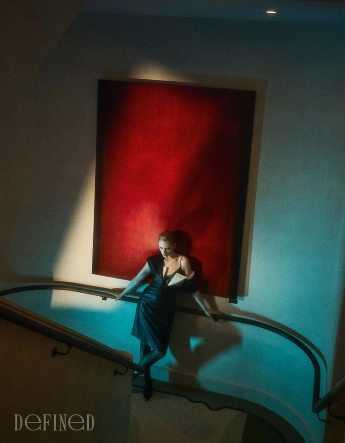 Actor Emma Laird leaning against a curved staircase railing in a dark dress before a red canvas in DEFINED Magazine.