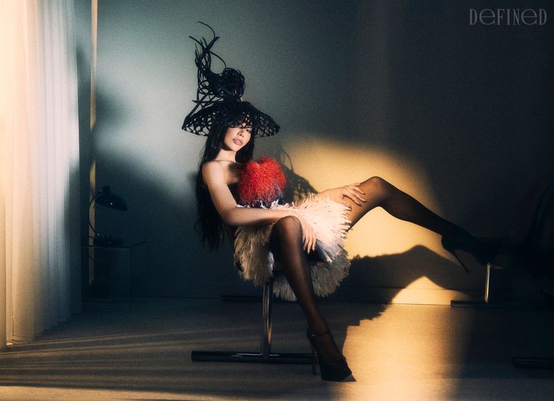 Munroe Bergdorf seated in a sculptural black mesh headpiece, feathered mini dress, and high heels under directional studio spotlight.