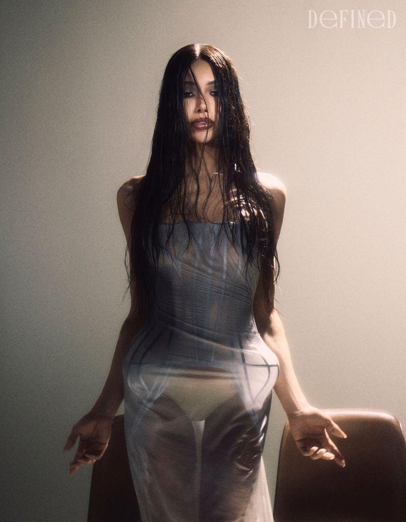 Model Munroe Bergdorf in a sheer grey dress with long dark wet hair standing against a plain background