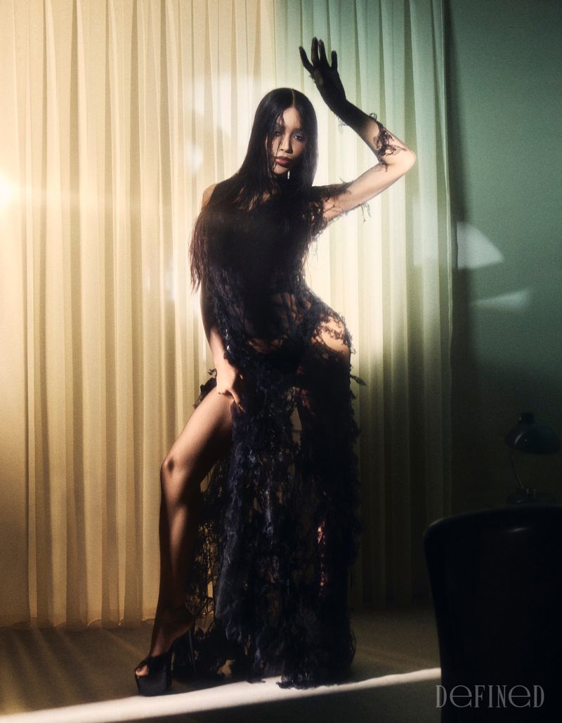 Munroe Bergdorf posing in a sheer textured black gown with a high leg slit and platform heels against backlit curtains.
