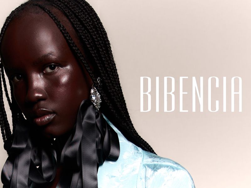 Close-up portrait of a Black female model with braided hair wearing black ribbon collar and light blue jacket against neutral backdrop.