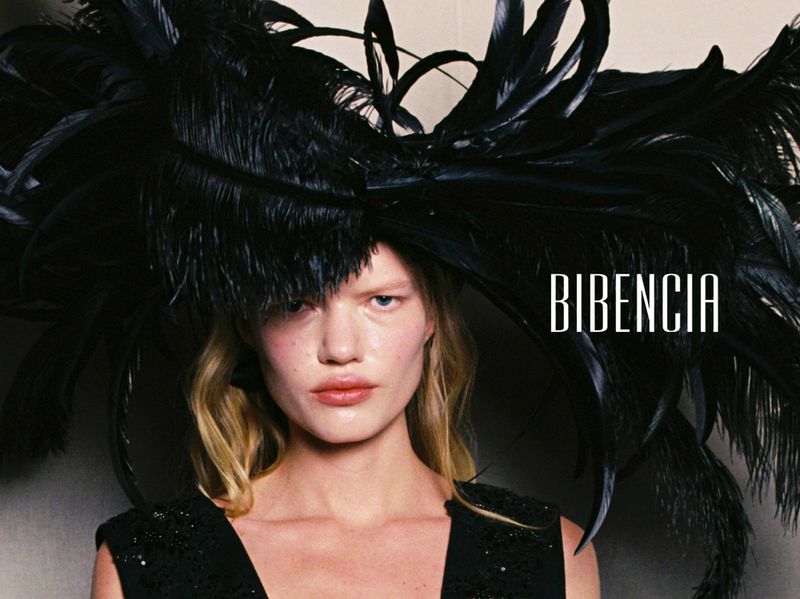 Model wearing an oversized black feather headpiece and embellished black sleeveless dress against a studio background.