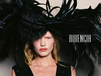 Model wearing an oversized black feather headpiece and embellished black sleeveless dress against a studio background.