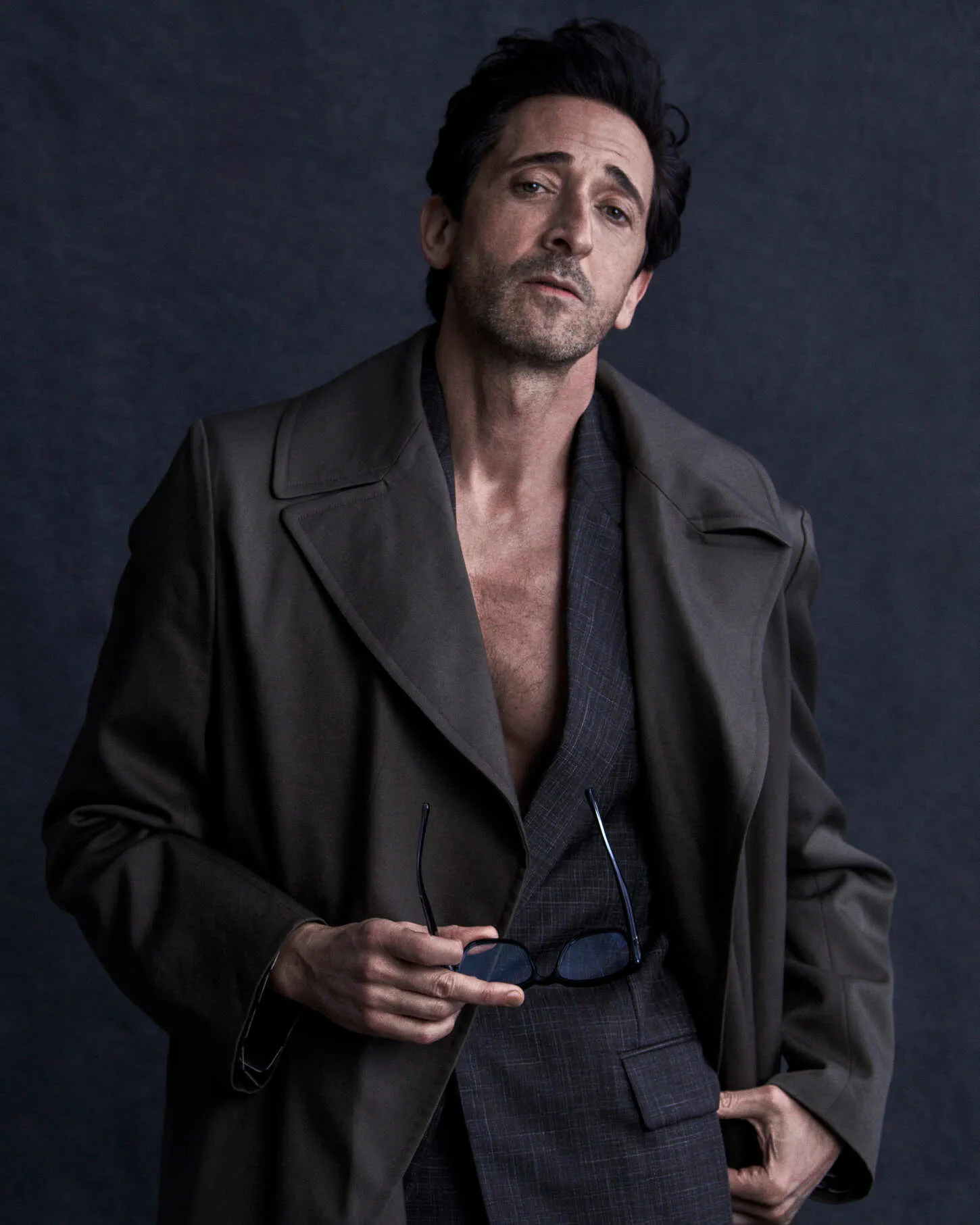 Actor Adrien Brody holding eyeglasses while wearing an open olive coat and charcoal suit jacket against a dark backdrop.