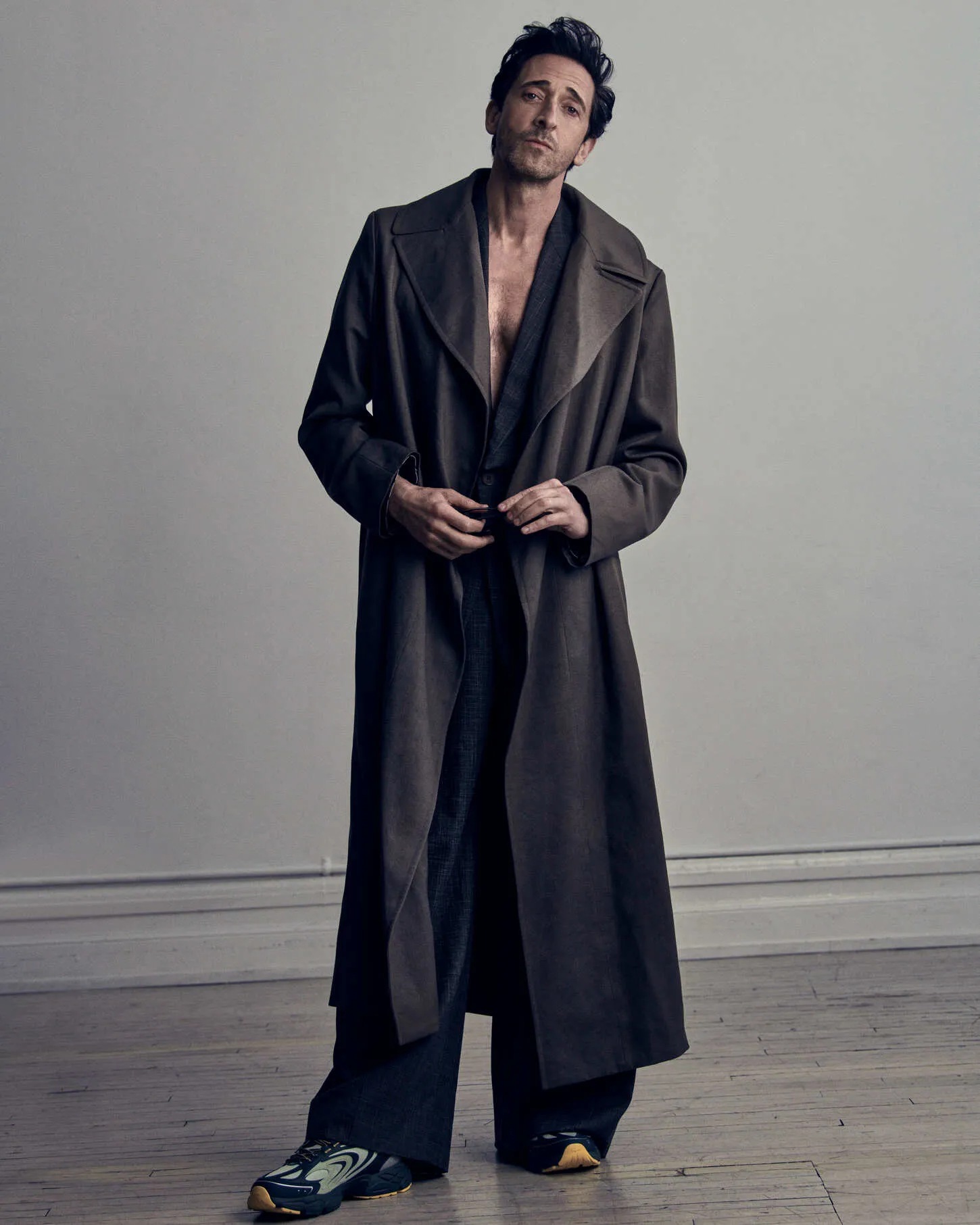 Actor Adrien Brody wearing a long brown coat and sneakers against a plain grey background