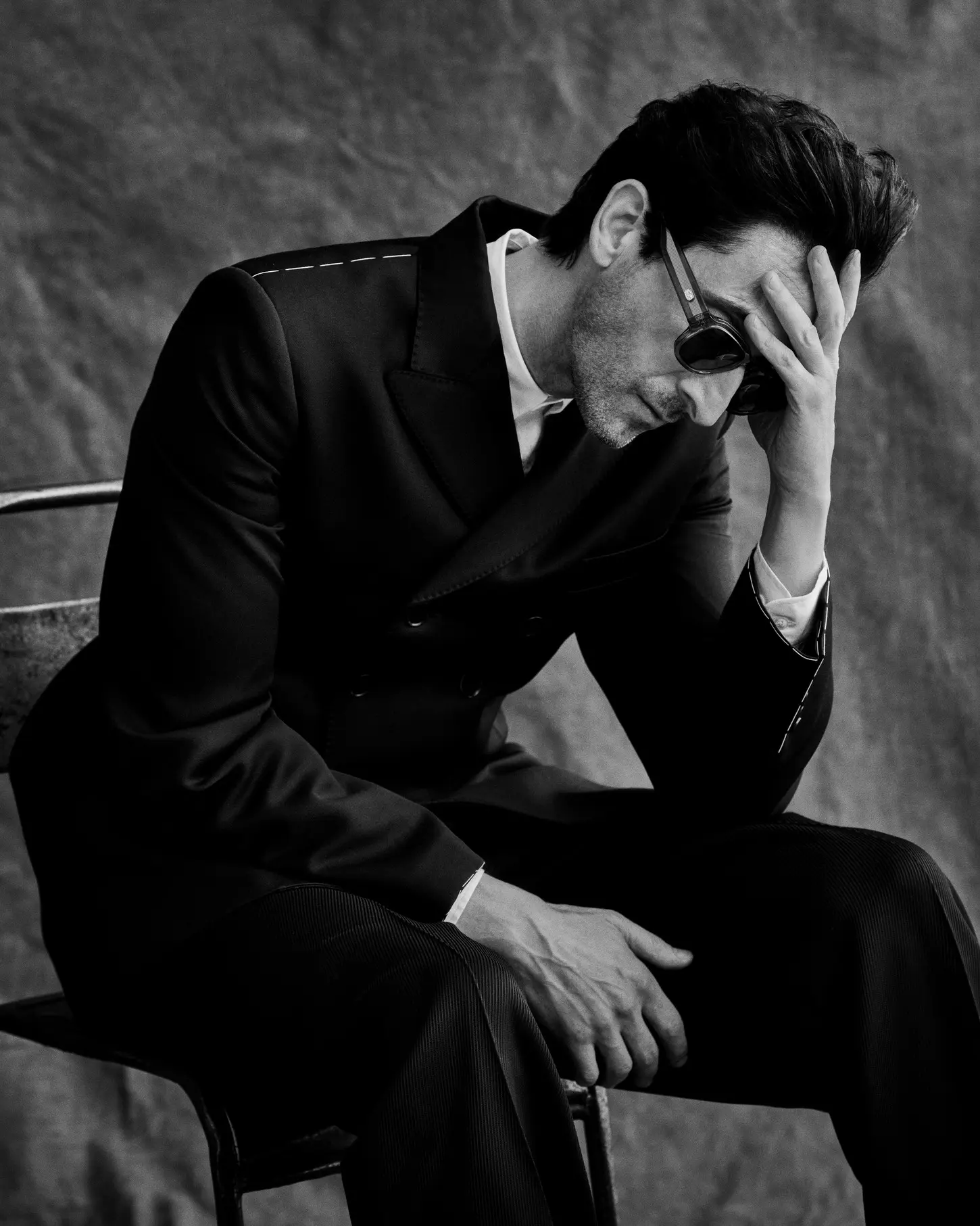 Actor Adrien Brody seated in a dark double-breasted suit and sunglasses with his hand to his head against a textured backdrop