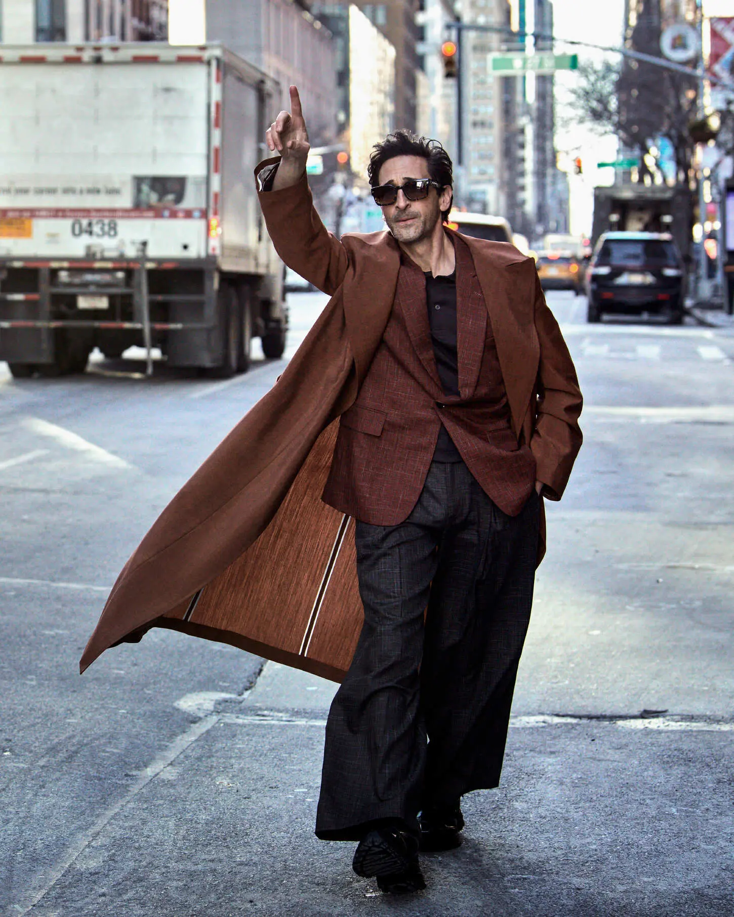 Actor Adrien Brody walks down an urban street raising his hand, dressed in a sweeping brown overcoat, blazer, and wide-leg trousers.