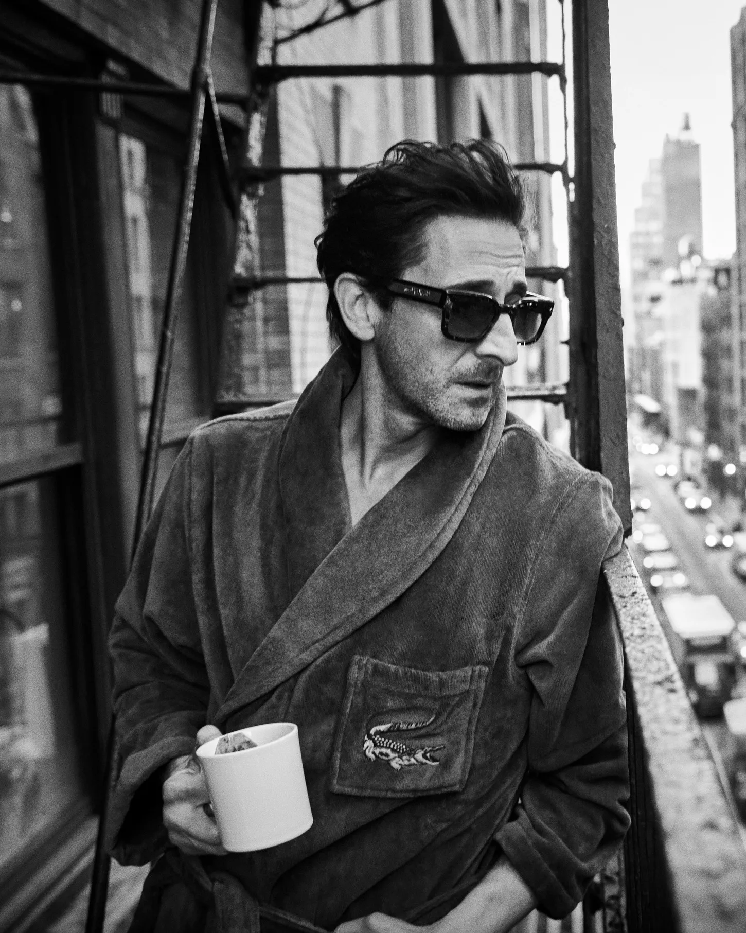 Actor Adrien Brody wearing a plush bathrobe and dark sunglasses while holding a white coffee mug on a city fire escape.