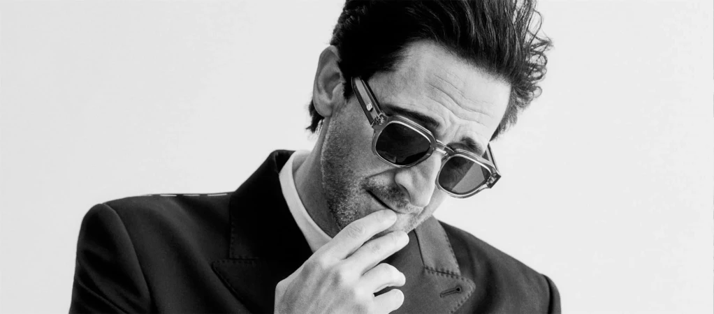 Adrien Brody wearing dark sunglasses and a black suit jacket while touching his chin against a bright white background.