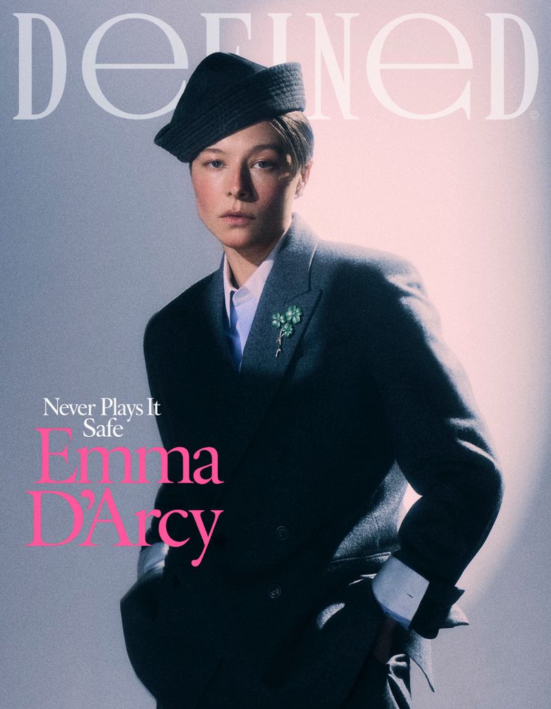 Emma D'Arcy wearing a dark tailored blazer and structured cap against a grey studio background for a magazine cover