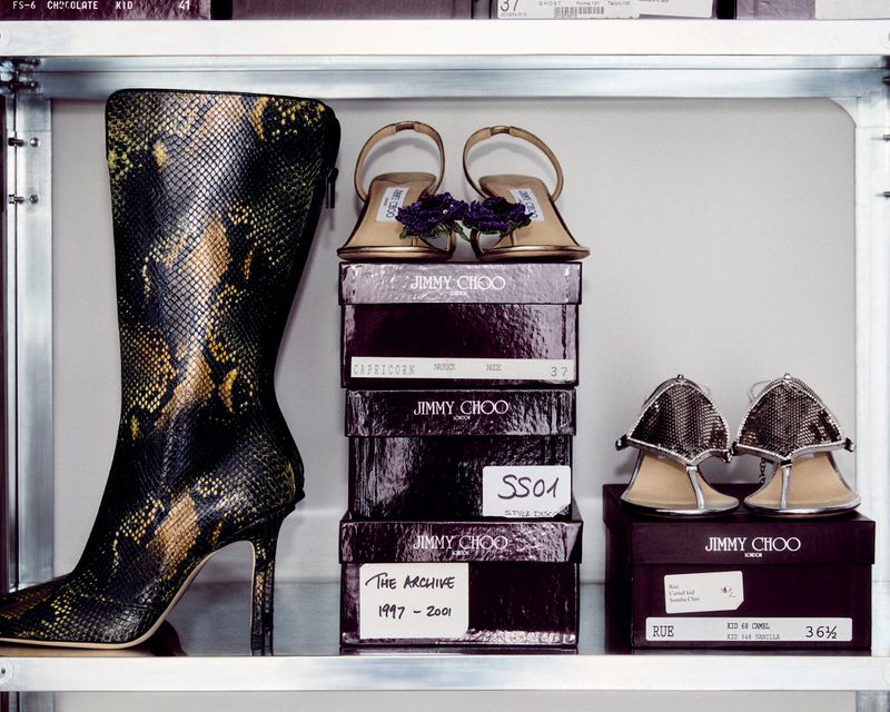 High angle shot of Jimmy Choo stiletto heels and a snakeskin boot on shelves, with product boxes, in a high-key setting.